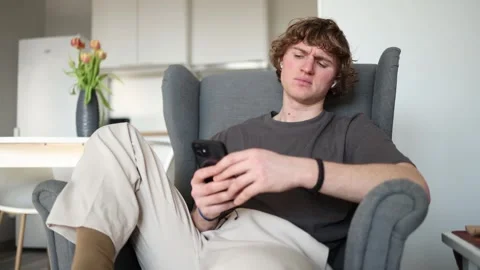 A slightly puzzled expression on a young man sitting in an armchair with a phone Stock Footage 302633827