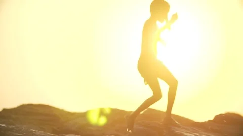 A slim boy climbs the rocky beach and dives in ocean at sunset Stock Footage 89269320