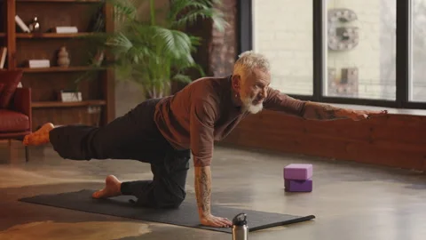 Slim silver-haired man practices tricky kitty yoga pose at home, active hobby Vídeo Stock 343507296