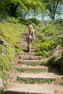Slim young blonde girl in a brown sundress climbs up the stairs in the park. Stock Photos