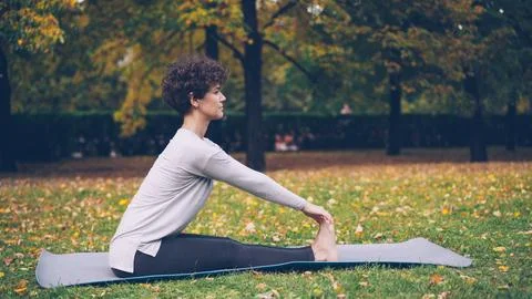 Slim young lady is doing sequence of yoga asanas sitting on mat in park alone on Foto stock