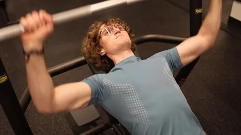 A slim young man, a student, is bench pressing in the gym. He is focused on his  Stock Footage 301677266