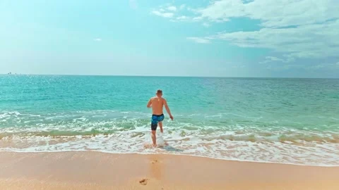 Slow-motion back view tracking shot of young man running into the sea Stock Footage 190022287