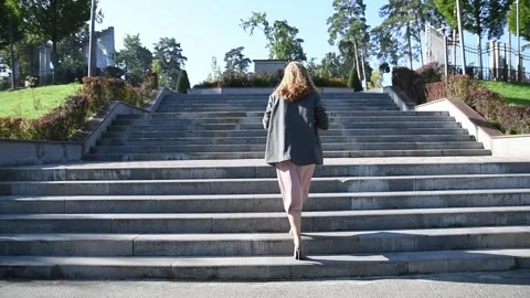 Slow motion back view of a young blonde woman walking up stairs in a park. 库存影片 333070714