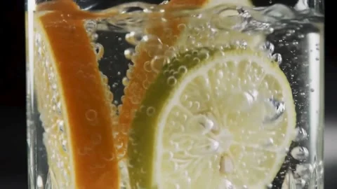 Slow-motion close-up of citrus fruit slices falling into sparkling water with bu Stock Footage 315397622