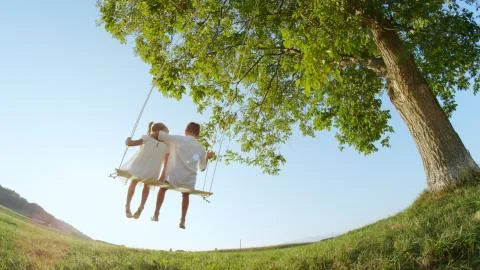 SLOW MOTION LOW ANGLE: Young boy hugging a little girl swinging on wooden swing Stock Photos