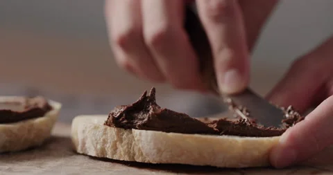 Slow motion man spreading chocolate nut spread on ciabatta slice on olive board Video stock 163599976