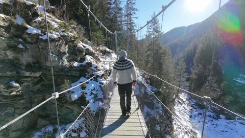 Slow motion man Walking Across Suspension Bridge Over River in Snowy Alps Stock Footage 326180531