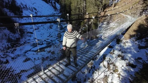 Slow motion man Walking Across Suspension Bridge Over River in Snowy Alps Stock Footage 326180610