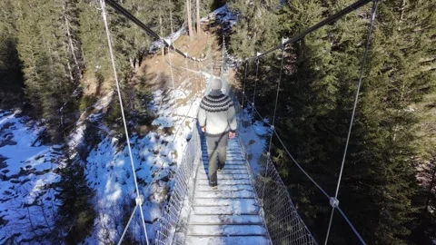 Slow motion man Walking Across Suspension Bridge Over River in Snowy Alps Stock Footage 326180649