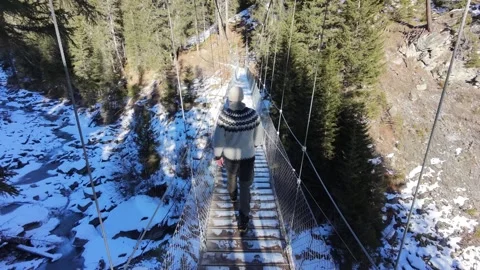 Slow motion man Walking Across Suspension Bridge Over River in Snowy Alps Stock Footage 326180663