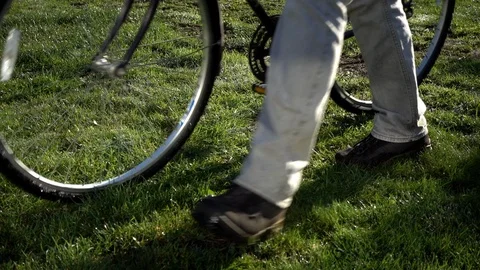 Slow motion – Man walking over wet grass pushing a bicycle, with long shadows. Stock Footage 98487010