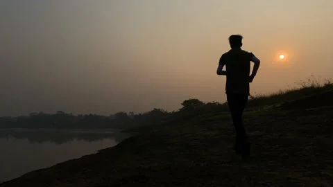 Slow motion rear view of young man jogging towards sun by river, hazy Stock Footage 233714852