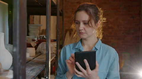Slow motion of serious girl using tablet in pottery workshop looking at ceramics Stock Footage 140774334