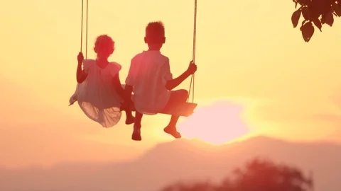 SLOW MOTION SILHOUETTE: Two happy kids swinging on wooden swing at golden sunset Stock Footage 82728062