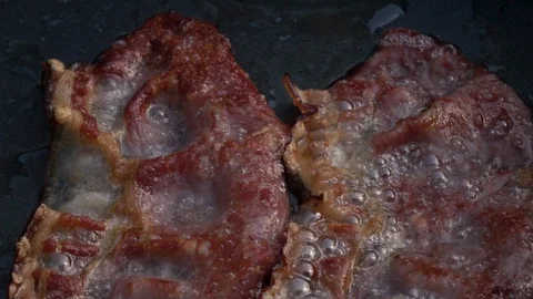Slow motion of sizzling sliced crispy wavy bacon on dark gray fry pan close up. Stock Footage 99850907