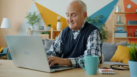 Slow motion of smiling senior man using laptop computer typing at desk in Stock Footage 128436842