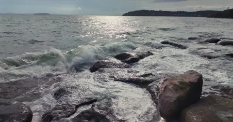 In slow motion, a sunlit wave explodes against dark rocks on the shore. Stock Footage 312566061