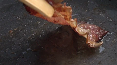 Slow motion of taking sizzling sliced crispy bacon away from dark gray fry pan Stock Footage 99851908