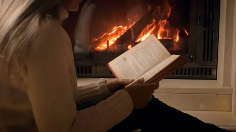 Slow motion video of young woman sitting at fireplace and reading book at night Stock Footage