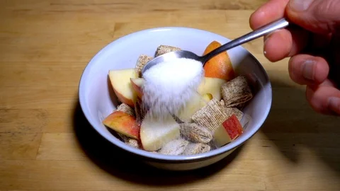 Slow motion: White sugar being sprinkled onto a bowl of wheat and apple cereal. Video stock 87330907