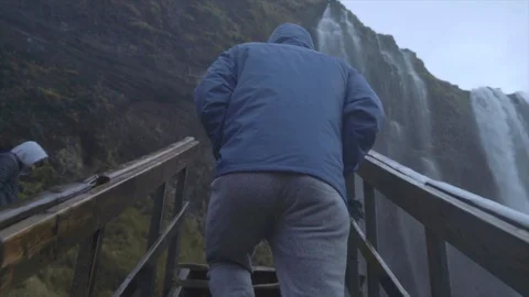 SLOW MOTION - Young man walking up steps toward Waterfall - Rainy Cloudy Day Stock Footage 129458300