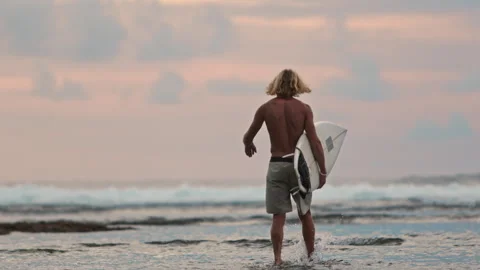 Slow motion: young surfer walking into water to surf breaking waves at soft pink Video stock 243818501