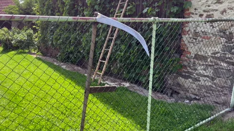 Slow pan reveals a full scythe hanging on a chain link fence in a green Stock Footage 327512840