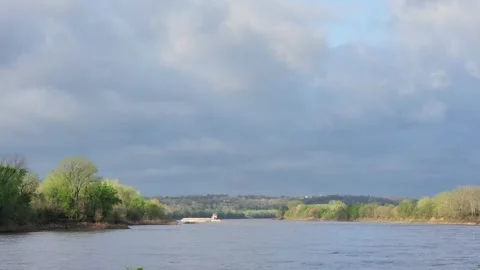 Slow river barge backing toward shoreline under dramatic sky Stock Footage 332884660