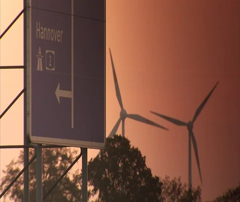 Slowly turning wind turbines next to road sign for Hannover, orange sky Stock Footage 86743261