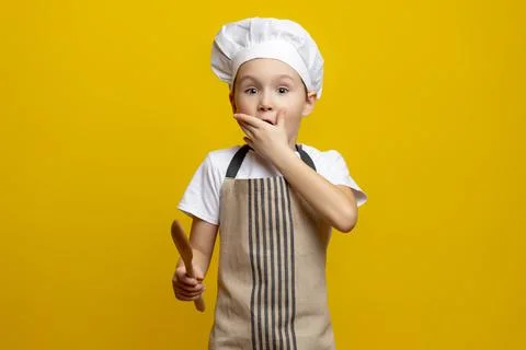 Small 6-7 year old boy holding a wooden spoon with an expression of surprise on Stock Photos