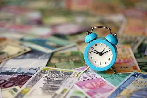 Small alarm clock on many banknotes of different currency. Background of time Stock Photos
