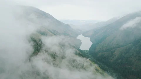 Small alpine lake surrounded by mountains appear from fog. Aerial flight Video stock 234490566