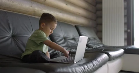 Small and cute boy learns to type on a laptop keyboard while sitting on a sofa Video stock 197638599