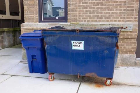 Small blue dented dumpster on wheels on a sidewalk next to a blue trash can Stock Photos