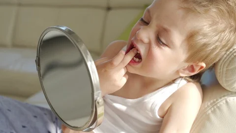 A small boy with blond hair with mirror opening his mouth to show where he lost Видео 107928161