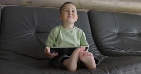Small boy in a bright room is playing with a digital tablet while sitting on the Stock Footage 197441772