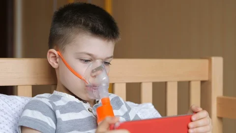 Small boy does therapeutic inhalation using a nebulizer and playing with a Video stock 126804872