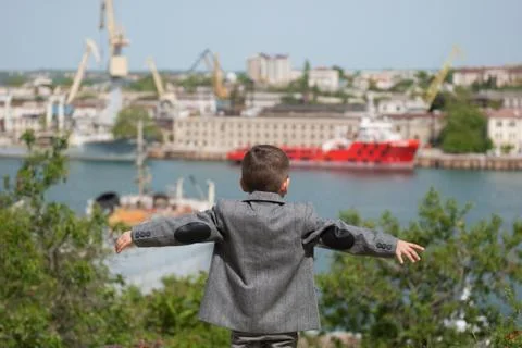 Small boy in a jacket stands against the backdrop of a seaport with his hands Foto stock