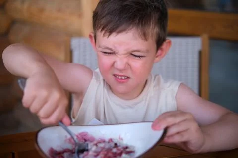 Small boy pulling a funny grimace face while eating food with a spoon in a bo Фото