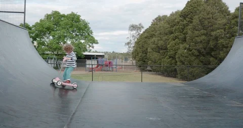 A small boy rides a pink three wheeled scooter on a skateboard half pipe ramp Stock-Footage 117358166