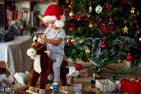 Small boy in Santa hat riding wooden rocking horse in front of christmas tree Stock Photos