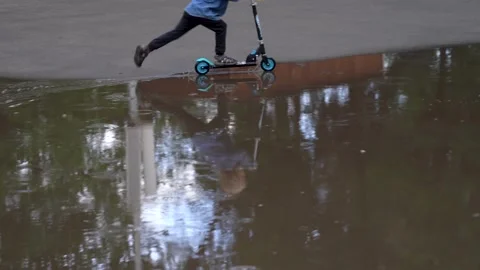 A small boy on a scooter rides through a large puddle in the rain Video stock 137006731
