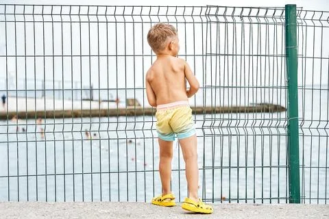 Small boy stands on parapet looking at seascape behind fence Foto stock