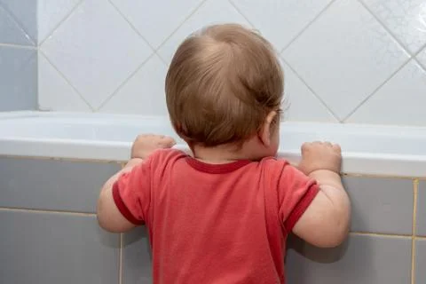 A small child in the bathroom looking into the bath, holding the edge Fotos de archivo