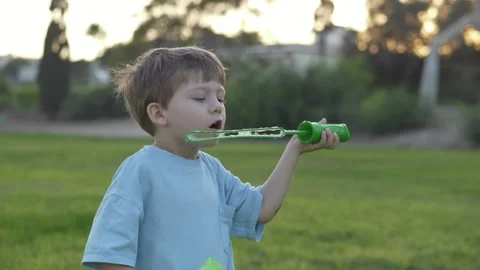 Small child carefully blows bubbles on the grass during sunny day. His serious Stock Footage 317318888