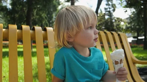 A small child eats ice cream cone sitting on a yellow bench in the park Stock Footage 213580903