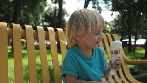 A small child eats ice cream cone sitting on a yellow bench in the park Stock Footage 213581034