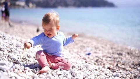 Small child hits pebbles with a stick while sitting on the beach by the sea Stock Footage 274500323