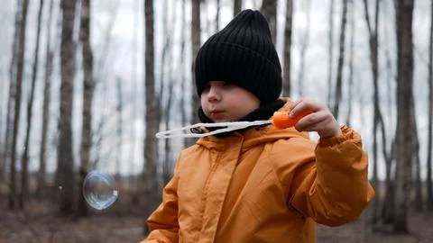 A small child in an orange jacket blows soap bubbles in the forest Stock Photos